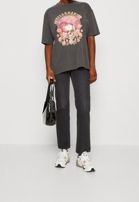 Persona con vaqueros negros desgastados y una camiseta gris oscuro de gran tamaño con un gráfico de calavera y flores, sosteniendo un bolso negro brillante, usando zapatillas blancas.
