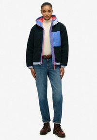 Jonge vrouw in een zwart en blauwe fleecejas met een capuchon, beige coltrui, blauwe jeans, een bruine riem en bruine veterschoenen, staand tegen een witte achtergrond.