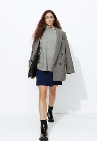 Blazer gris oversize, pull col roulé en maille grise, jupe texturée marine, bottines noires, tenant un sac à main noir.