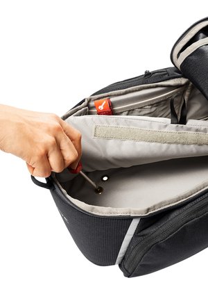 Zaino nero con interno grigio, dotato di cerniera, più tasche e una mano che tiene uno strumento. Include una tracolla in tessuto e un'etichetta di branding.