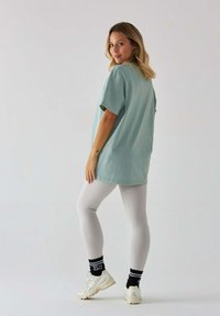 Jasnozielona, oversize t-shirt z krótkim rękawem, zestawiona z dopasowanymi szarymi legginsami. Białe sneakersy z czarno-białymi paskami na skarpetkach. Proste tło.