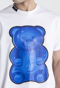 T-shirt bianca in cotone con una grande grafica di un orsetto gommoso blu con texture lucida e contorno nero. Piccola etichetta visibile sul colletto.