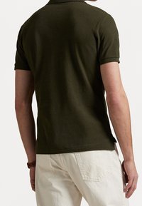 Man som bär en mörkgrön kortärmad pikétröja och ljusbeige byxor, visad bakifrån med en brun klocka på vänster handled.