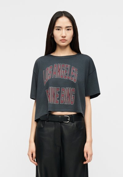 Junge Frau mit langen dunklen Haaren, die ein kurzes dunkelgraues T-Shirt mit der Aufschrift "Los Angeles Mine Bing" und schwarze lederne Hosen mit Gürtel trägt.