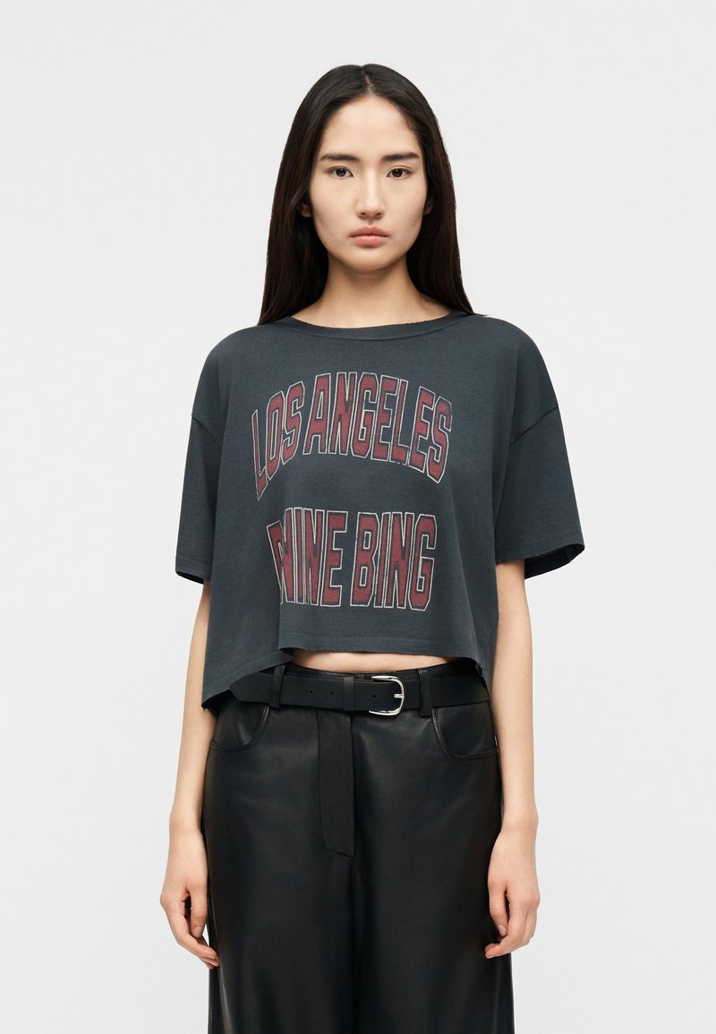 Νεαρή γυναίκα με μακριά σκούρα μαλλιά, που φοράει ένα κοντό σκούρο γκρι T-shirt με την επιγραφή "Los Angeles Mine Bing" και μαύρο δερμάτινο παντελόνι με ζώνη.