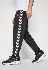 Čierne športové nohavice majú biely chevron pás pozdĺž boku, s úzkym strihom a elastickým pásom. Nosia sa s čiernymi teniskami.