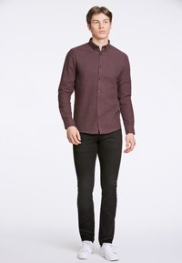 Camicia bordeaux a maniche lunghe con bottoni e tasca, abbinata a jeans neri slim-fit e sneaker bianche. Tessuto morbido e design aderente.