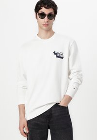 Hvid sweatshirt med rund hals, forsynet med et marineblåt "TOMMY JEANS" logo på venstre bryst. Kombineret med mørke denim jeans og solbriller.