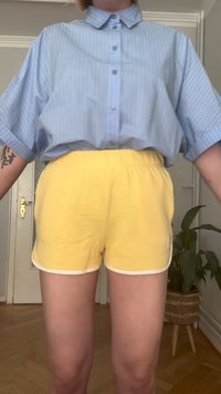 Camisa holgada a rayas azul claro con botones, combinada con pantalones cortos amarillos de cintura elástica con ribete blanco. Ambiente interior con plantas visibles.