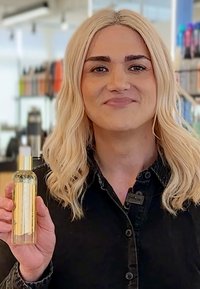 Bottiglia di vetro trasparente con una pompa dorata, riempita con un liquido giallo chiaro. Persona che la tiene, indossando una camicia nera con bottoni e capelli chiari.