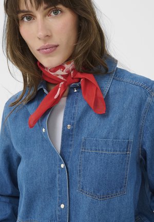 Vaaleaihoinen nainen, jolla on vihreät silmät ja punakuvioinen kaulahuivi, valkoinen paita ja sininen farkkutakki nappeineen.