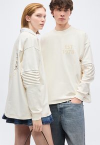 Νεαρή γυναίκα με εκρού φούτερ και τζιν φούστα κρατάει καφέ τσάντα, στέκεται δίπλα σε νεαρό άντρα με εκρού φούτερ και μπλε τζιν.