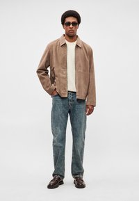 Barna szarvasbőr dzseki galléros nyakkivágással, krémszínű ing fölött viselve. Kék farmerrel és sötétbarna cipővel párosítva. Egyszerű kialakítás, laza fazon.