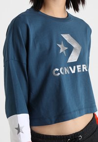 Oříznutá modrá bavlněná mikina se stříbrným textem "CONVERSE" a hvězdicovým logem, s rukávy v barevném provedení bílé a červené. Měkká textura.