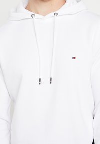 Witte hoodie met een voorzak, trekkoorden met zilveren aglets en een klein geborduurd logo op de borst in rood, wit en blauw.