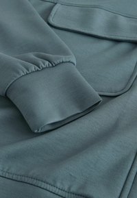 Blue-green fabric with a smooth texture, featuring a cuffed sleeve and a patch pocket. The stitching is even, highlighting the garment's quality.