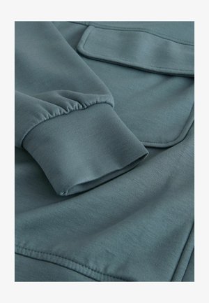 Tkanina v modro-zelené barvě s hladkou texturou, se s volným rukávem a náprsní kapsou. Švy jsou rovnoměrné, což podtrhuje kvalitu oděvu.