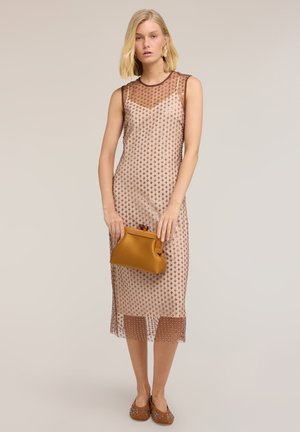 Kvinde i ærmeløs, knælængde, gennemsigtig beige kjole med brune polkaprikker, holder en sennepsfarvet clutch og har brune pyntede flade sko på.