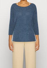 Maglione blu con texture, scollatura ampia e maniche a tre quarti, abbinato a pantaloni larghi color crema.