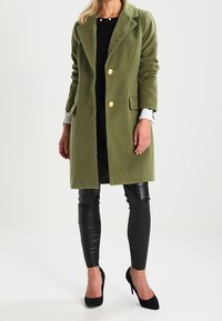 Olivgrön lång kappa med slagkrage, två gyllene knappar, sidofickor och en mjuk textur. Kombinerad med åtsittande svarta läderbyxor.