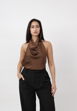 Vrouw met lang donker haar draagt een mouwloze bruine satin top en zwart hoog getailleerde broek, staand tegen een effen lichte achtergrond.