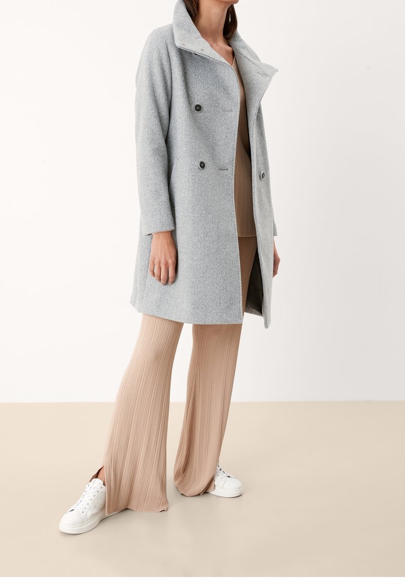 Cappotto grigio in misto lana con design doppiopetto, collo a revers e bottoni scuri. Abbinato a pantaloni beige a coste e sneakers bianche.