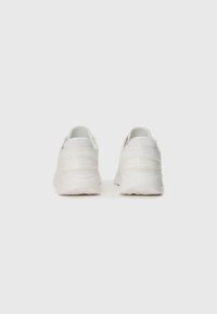White sneakers featuring a smooth synthetic upper, padded collar, and textured heel accents; rubber sole with patterned tread.