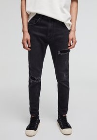 Vaqueros de mezclilla negra con corte ajustado, que presentan parches desgastados en las rodillas, un ligero desvanecimiento y bolsillos traseros; combinados con zapatillas negras.