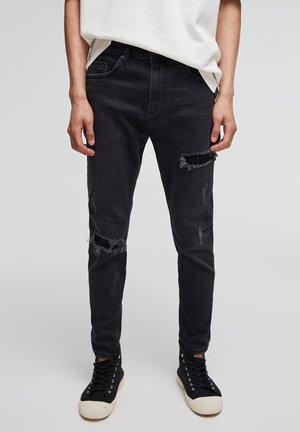 Vaqueros de mezclilla negra con corte ajustado, que presentan parches desgastados en las rodillas, un ligero desvanecimiento y bolsillos traseros; combinados con zapatillas negras.