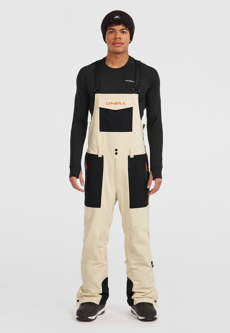 Mono de nieve beige con acentos negros, tirantes ajustables, bolsillos frontales y cremalleras. Diseñado para brindar calidez y movilidad en condiciones invernales.
