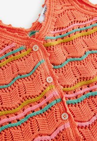 Stickad cardigan i stark orange med dekorativa vågmönster i grön, rosa och gul. Har runda knappar och öppet arbete-detaljer.