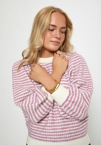Stickad tröja med rosa och vita ränder, med en ribbad rundhals. En person med guldfärgade armband och ringar på korsade armar.