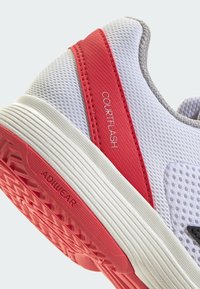 Chaussure de sport blanche avec des accents rouges, tige en mesh et semelle en caoutchouc texturé. Présente les marques "COURTFLASH" et "ADIWEAR" sur le côté.