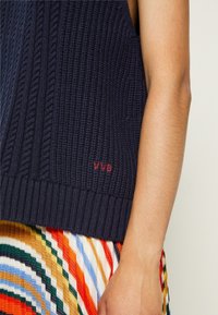 Primer plano de una persona que lleva un chaleco de punto azul marino con bordado rojo "VVB" y una falda de rayas multicolores.