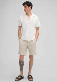 Weißes Kurzarm-Polohemd mit Kragen, kombiniert mit beigen gestreiften Shorts. Schwarze Sandalen und ein Bein-Tattoo sind sichtbar. Neutraler Hintergrund.