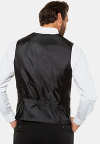 Chaleco de satén negro, con una textura suave, detalle de cinturilla en la espalda y una silueta estructurada, llevado sobre una camisa blanca.