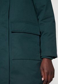 Mørkegrøn jakke med en tekstureret overflade, der har en stor forlommeså og ribbede manchetter. Stoffet er glat og vandafvisende.