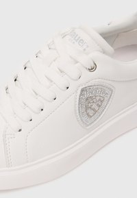 White low-top sneaker with white laces, silver eyelet, and a silver-embellished brand patch on the side.