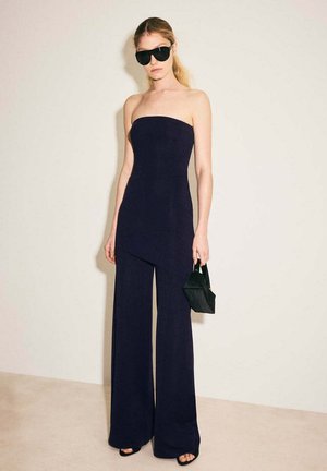 Mujer con mono negro sin tirantes, gafas de sol negras, sosteniendo un bolso pequeño negro, de pie frente a un fondo liso y claro.