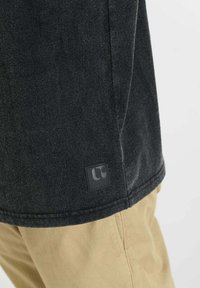 Zwart, T-shirt met korte mouwen en een gestructureerde afwerking, met een rechthoekige logopatch nabij de zoom. Gecombineerd met beige broek.