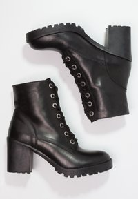 Botas de tobillo de cuero negro con un tacón grueso y suela robusta. Presentan ojales de metal y cordones en la parte delantera, textura suave y un diseño elegante.
