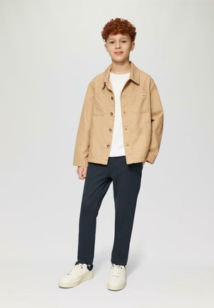 Lichtbruine knopenjacket over een wit shirt, gecombineerd met donkere broeken en witte sneakers. Gladde stof, ontspannen pasvorm en twee voorzakken.