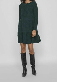 Femme portant une robe côtelée à manches longues couleur vert foncé et des bottes à talons noires montant jusqu'aux genoux, se tenant devant un fond gris clair uni.