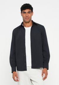 Camisa negra de botones con cuello clásico y mangas largas, llevada sobre una camiseta blanca. Tejido liso sin patrones o acentos visibles.