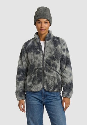 Femme portant une veste polaire grise à motifs avec des poches zippées, un jean bleu, une chemise blanche et un bonnet en tricot gris sur un fond uni.