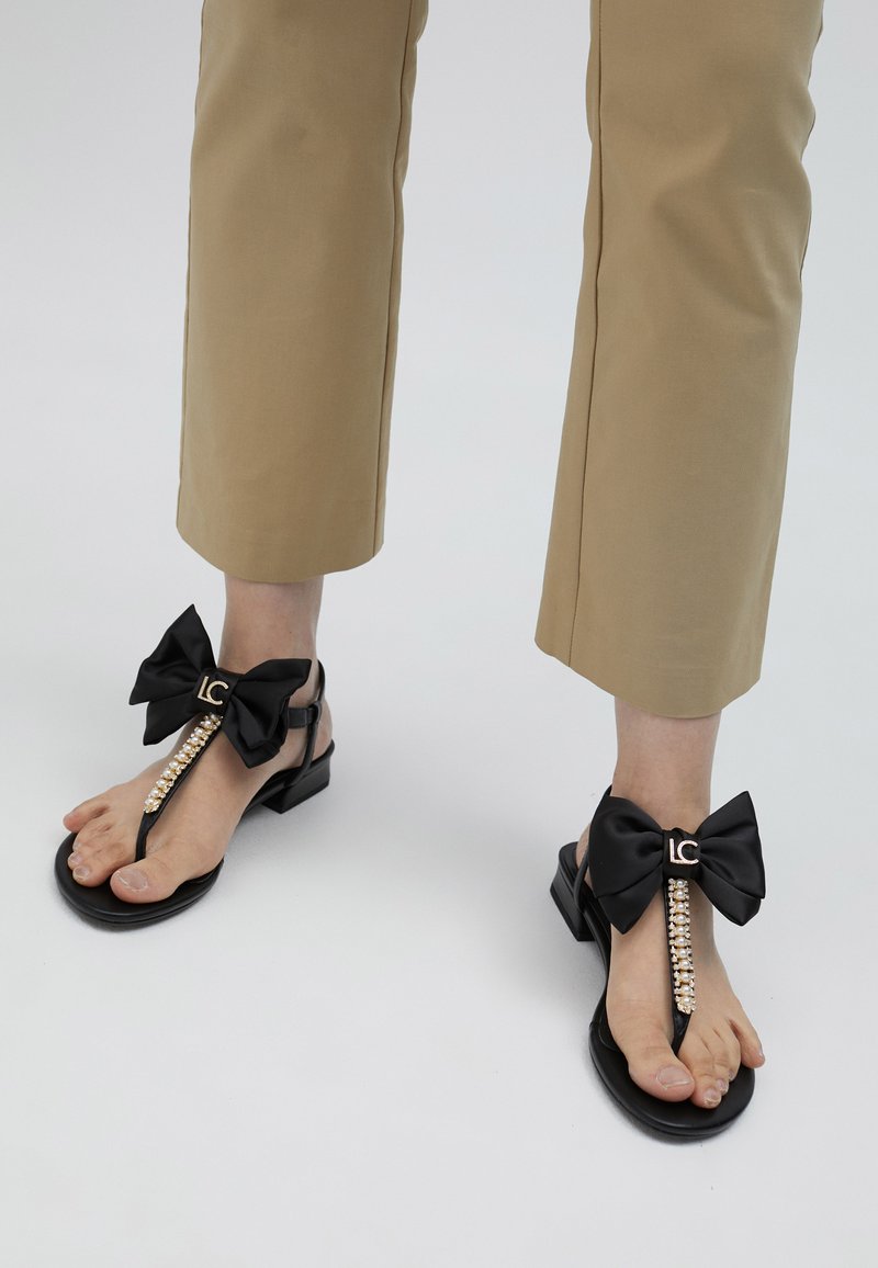 Black sandals with a satin bow detail, embellished with a pearl-like accent and a logo, feature a flat sole and minimalist design.