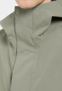 Hellegrüne Jacke mit hohem Kragen, die eine glatte Textur und kontrastierende Nähte entlang der Nähte aufweist. Minimalistisches Design.