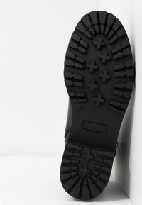 Semelle de botte en caoutchouc noir avec des crampons profonds et des motifs en forme de croix, taille 37, positionnée devant un fond blanc en coin.