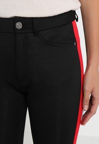 Schwarze Hosen mit glatter Textur, ausgestattet mit einem roten Seitenstreifen und einem Metallknopfverschluss. Das Design umfasst Vorderaschen und Gürtelschlaufen.