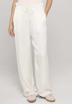 Pantalon blanc ample avec taille à cordon, porté avec des baskets rose clair et un haut rose clair, debout devant un fond neutre.